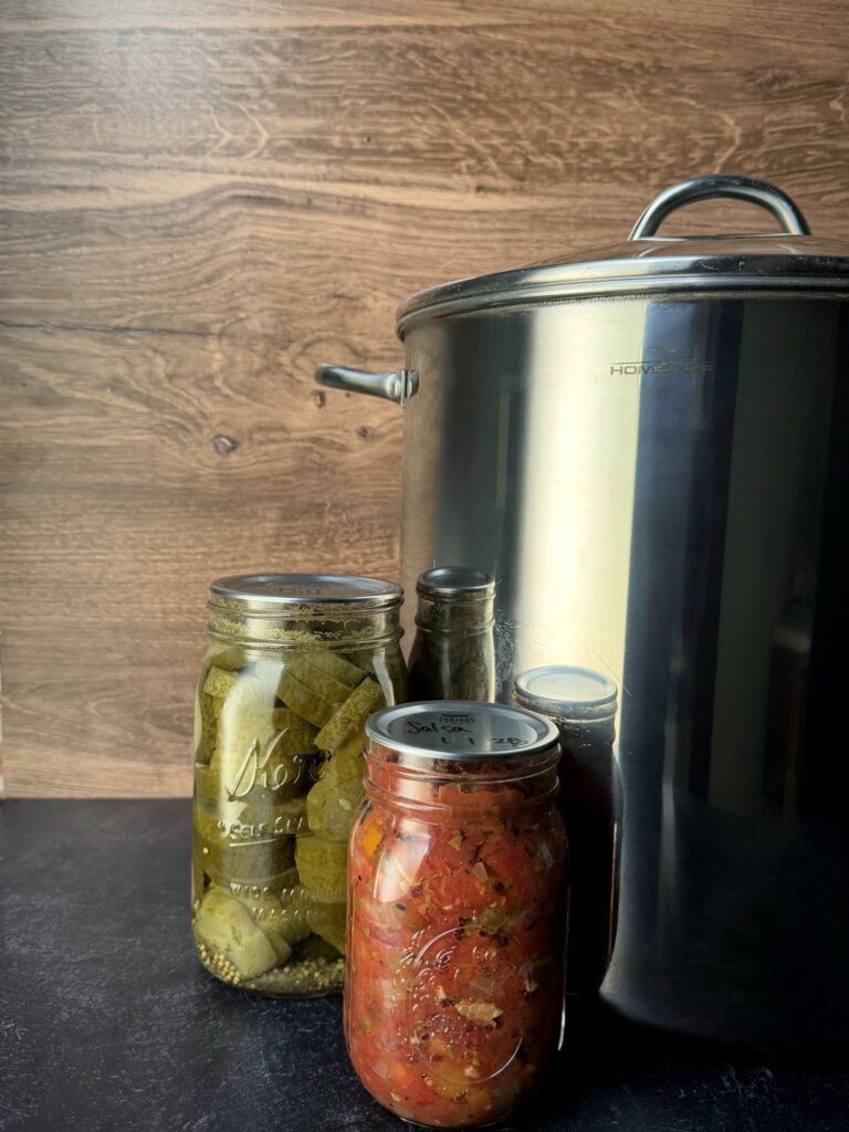 Stainless steel 20-quart stockpot with lid beside jars of home-canned pickles and salsa