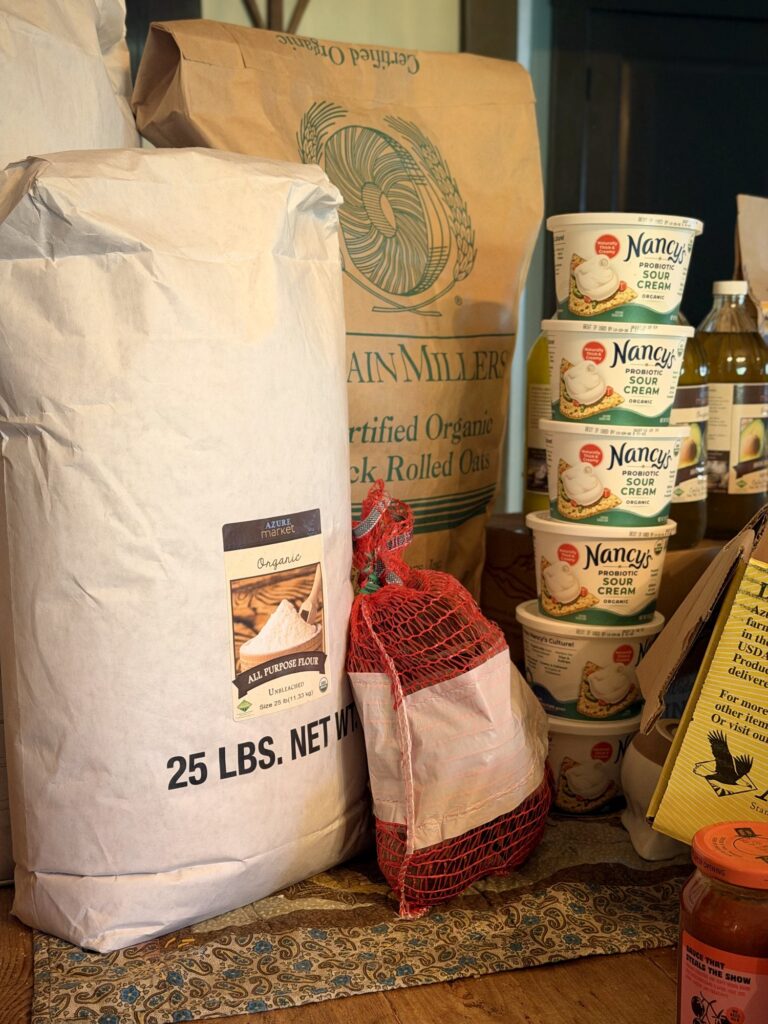 A 25-pound bag of organic all-purpose flour next to a large sack of rolled oats, a red mesh produce bag and a tall stack of sour cream tubs