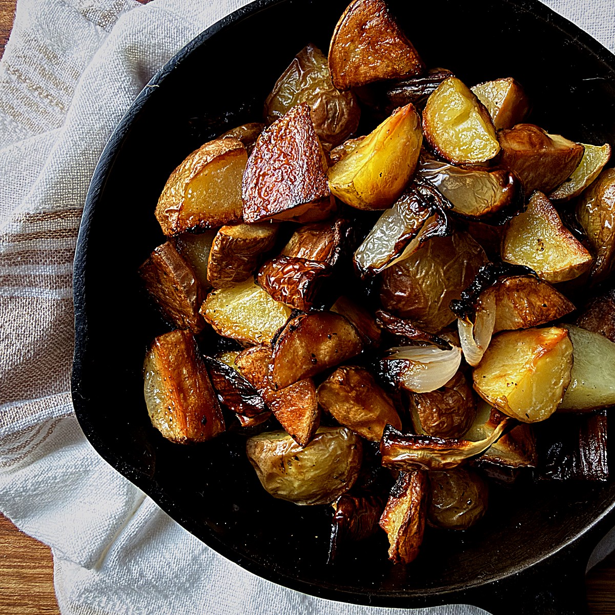 Crispy roasted potatoes and onions in a cast iron skillet on a striped kitchen towel.