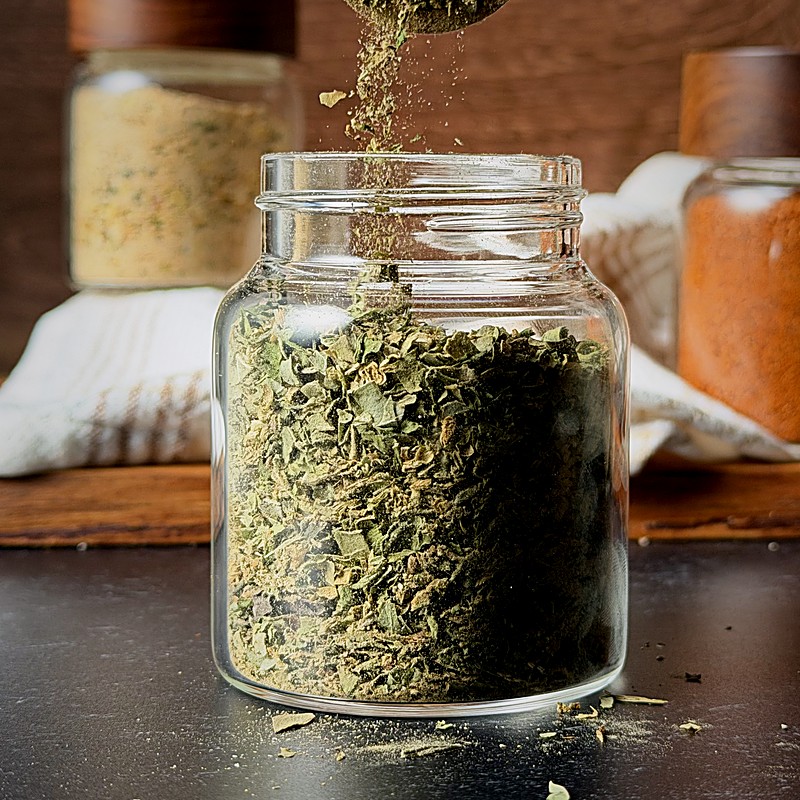 Dried green herbs being sprinkled into a glass spice jar.