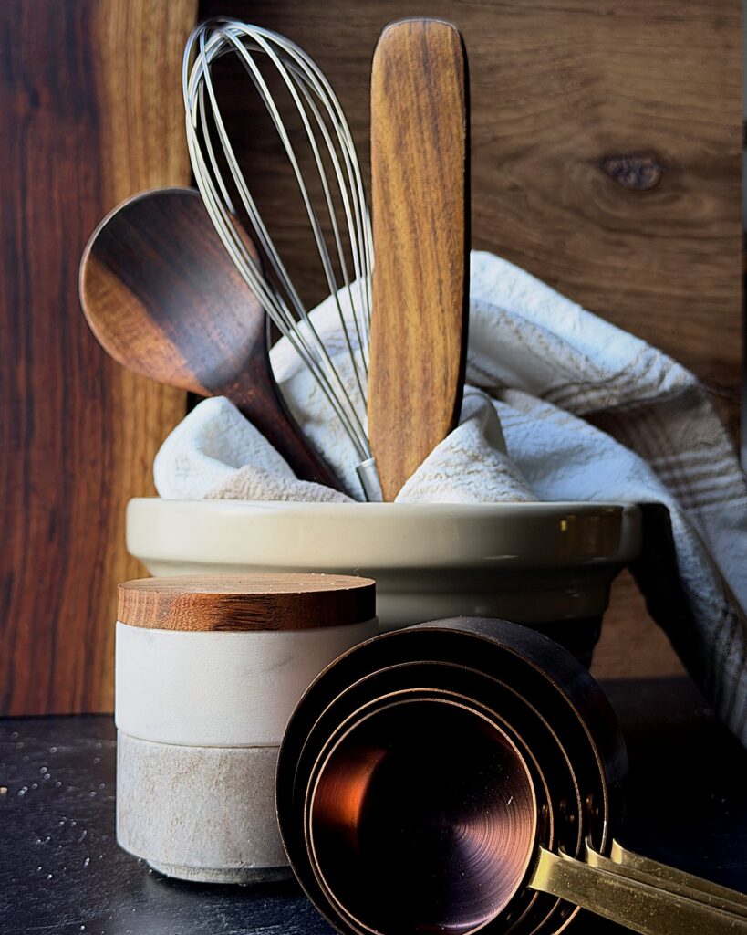 Stoneware utensil crock holding wooden spoons and a whisk, with copper measuring cups in front.