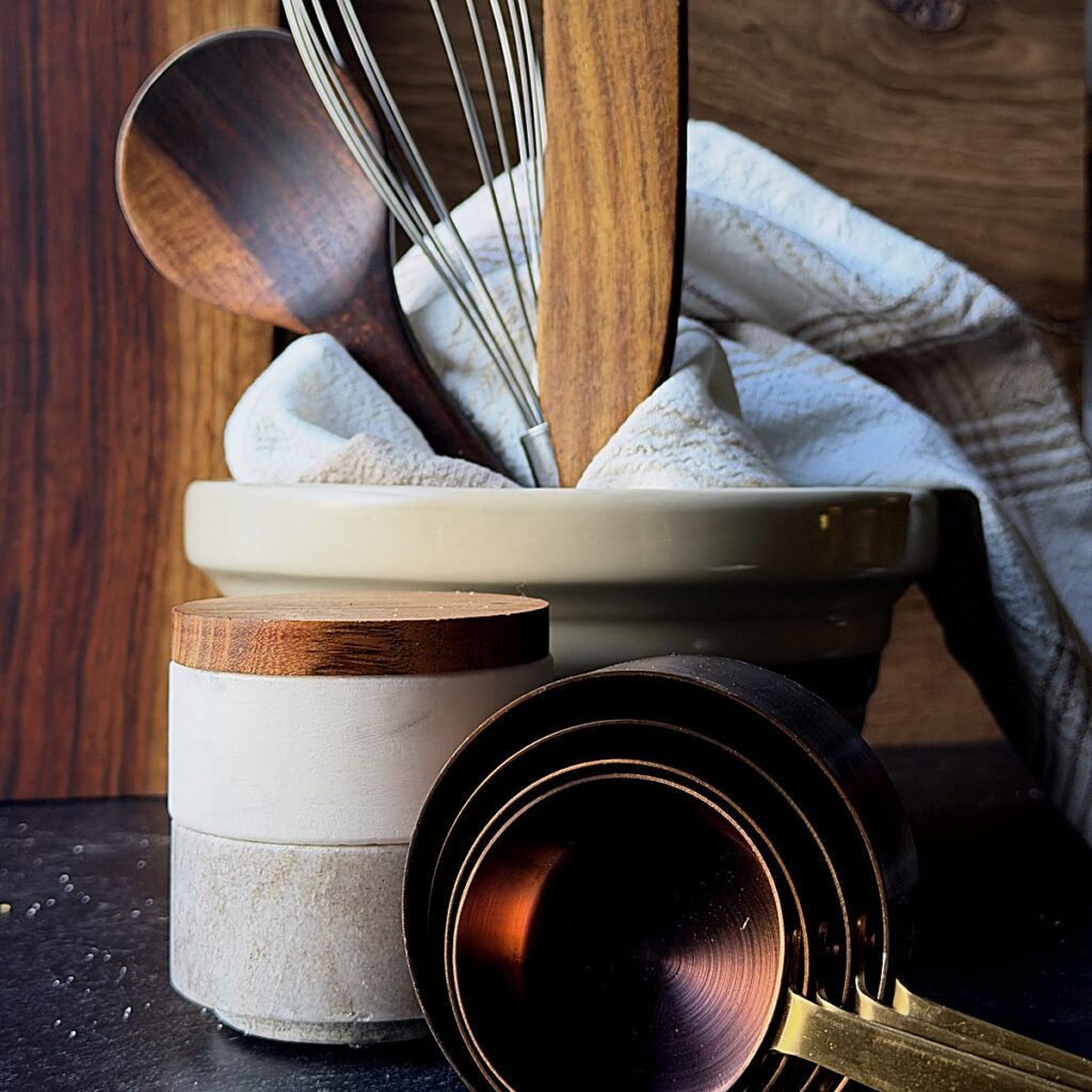 Wooden spoons and a whisk in a stoneware crock beside copper measuring cups.