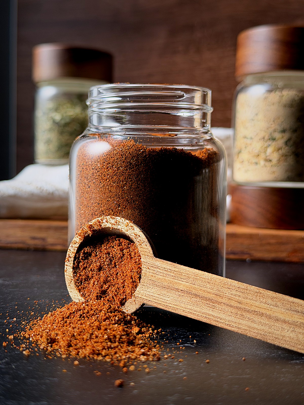Wooden scoop of red spice blend spilling onto a dark counter beside a glass jar.