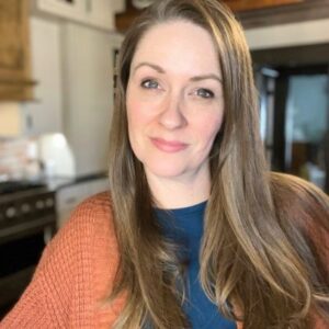 Headshot of a smiling woman in an orange sweater in a kitchen.