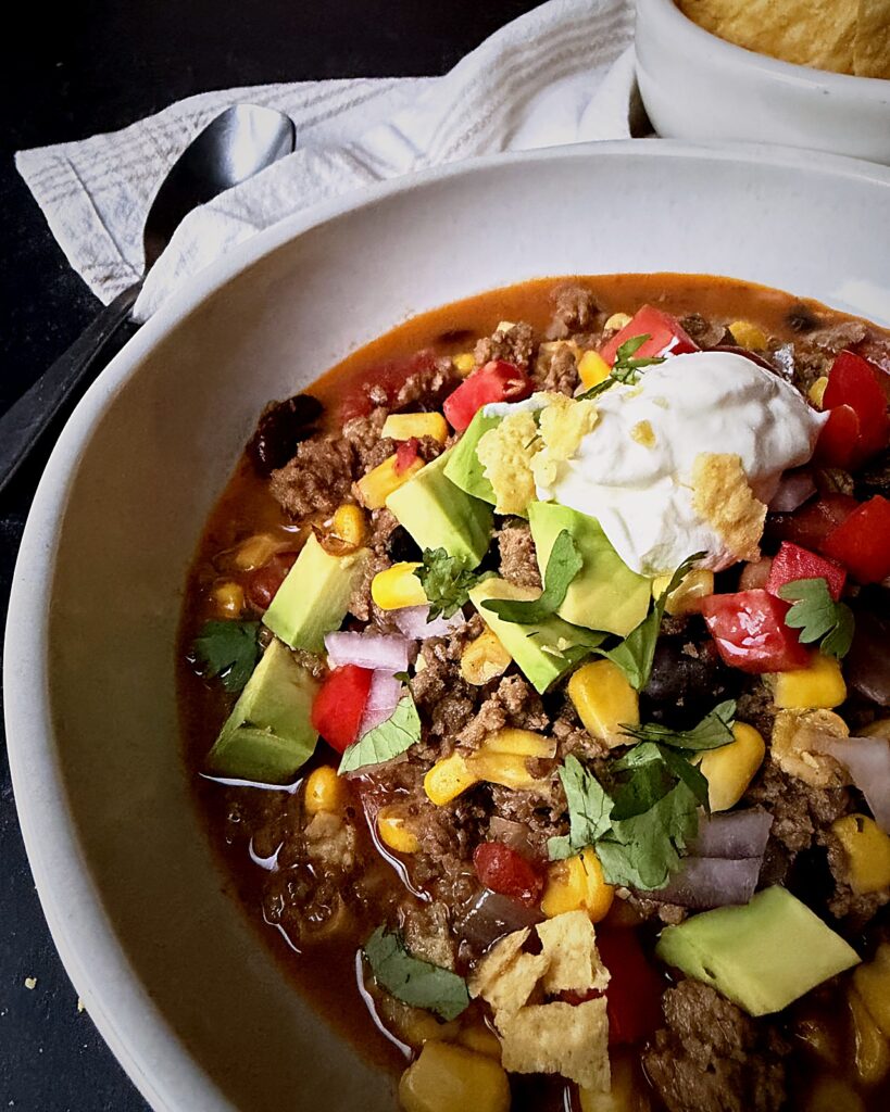 Bowl of taco soup with ground beef, avocado, and sour cream, with tortilla chips on the side.