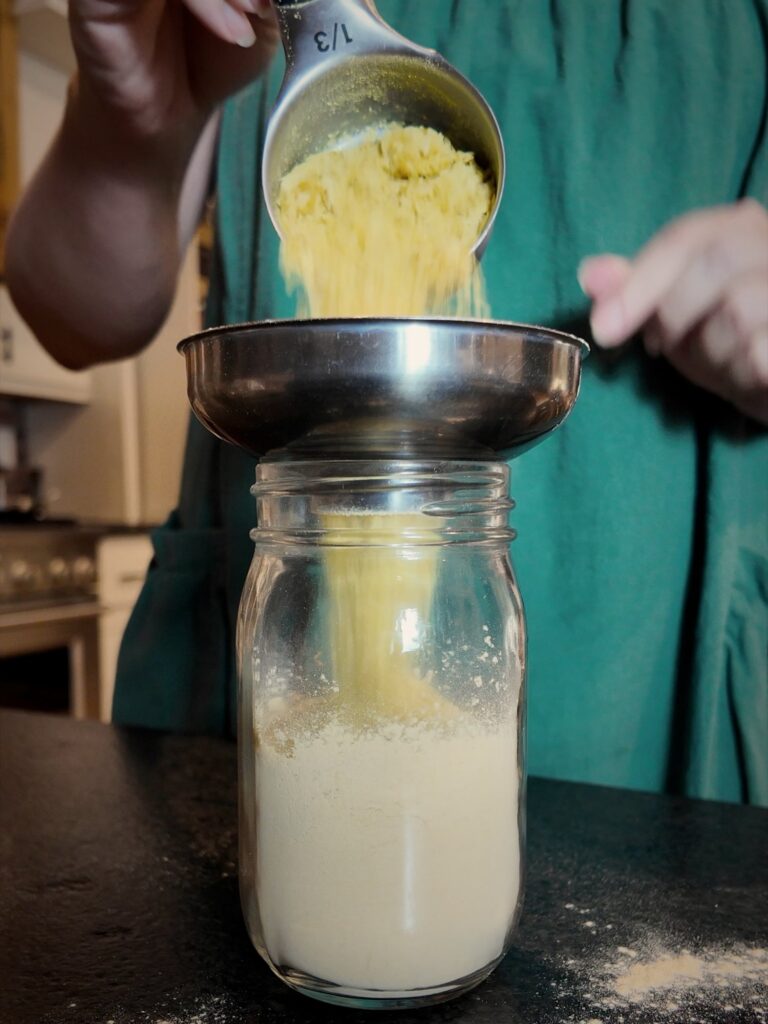 Nutritional yeast poured from a measuring cup through a stainless funnel into a jar of broth powder