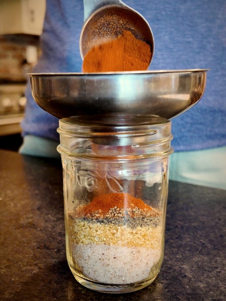Adding paprika through a canning funnel into a jar layered with burger seasoning ingredients