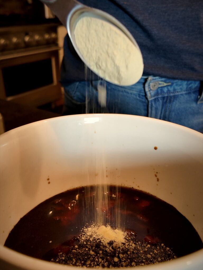 Garlic powder pouring from a measuring spoon into a soy sauce jerky marinade topped with cracked black pepper