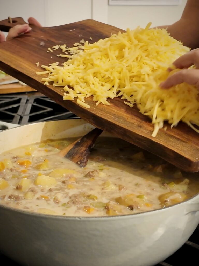 Shredded cheddar cheese sliding off a wooden cutting board into a pot of creamy cheeseburger soup.