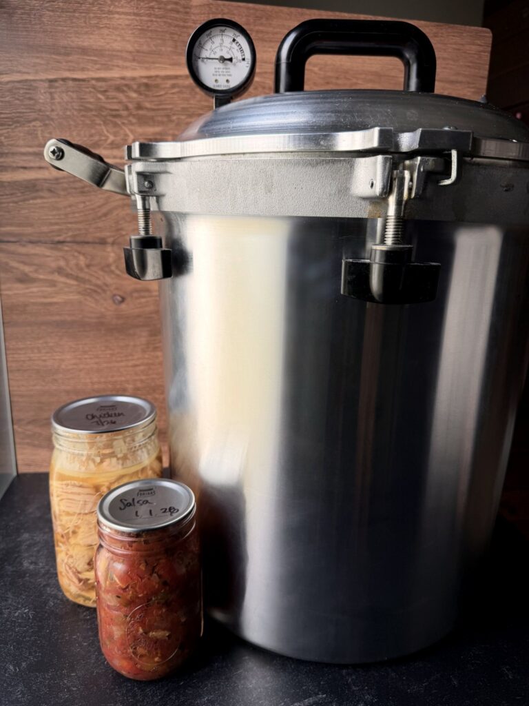 All-American pressure canner with a pressure gauge and clamp knobs on the lid, next to jars of home-canned food