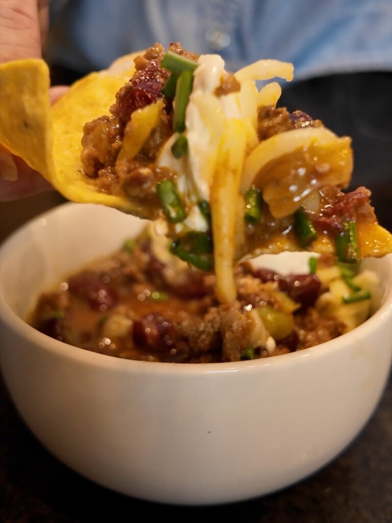 Chip scooping chili topped with cheese, sour cream and green onions from a white bowl