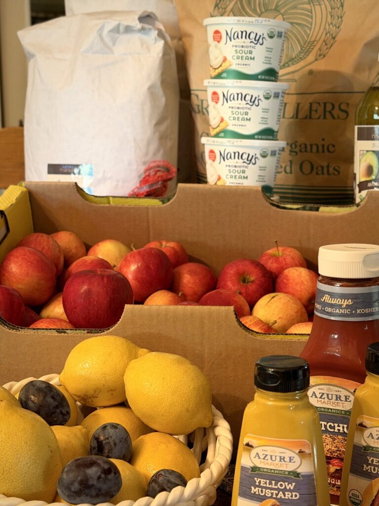 Cardboard boxes of red apples with a bowl of lemons and plums, organic yellow mustard and ketchup in front, and bulk flour, rolled oats and sour cream stacked behind