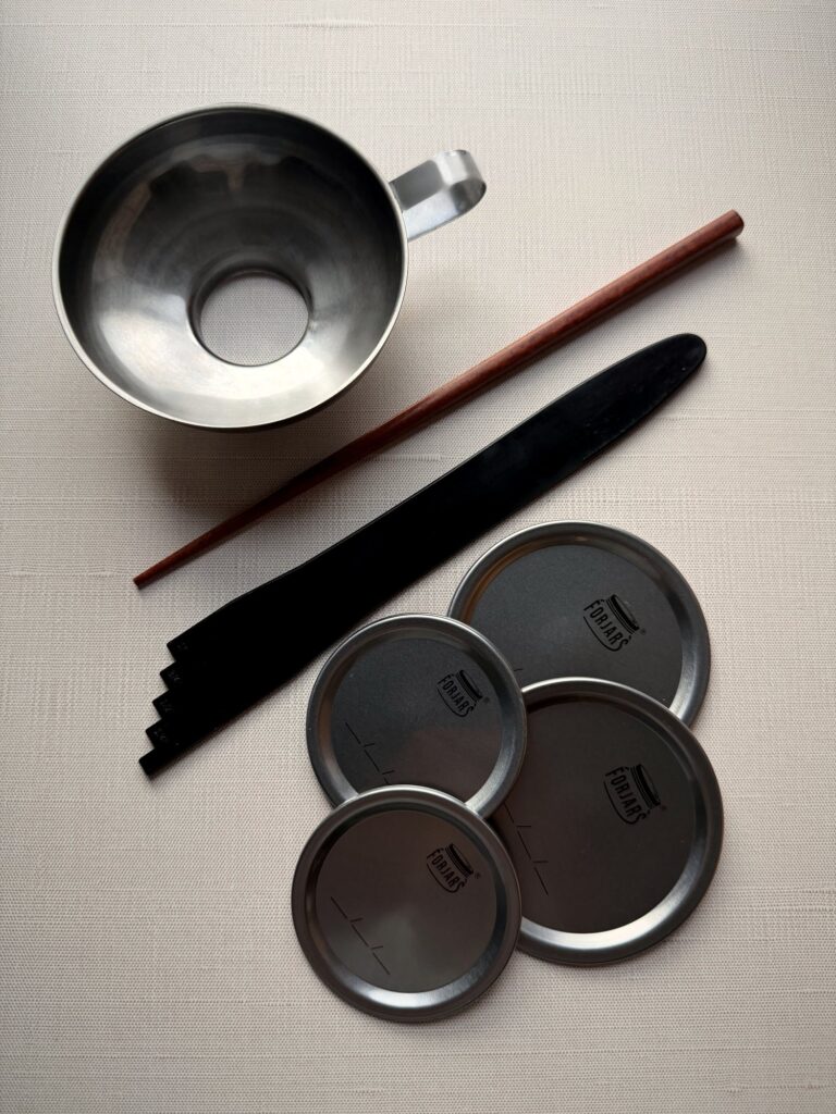 Stainless steel canning funnel, wooden bubble stick, black stepped headspace tool and four ForJars lids on white linen