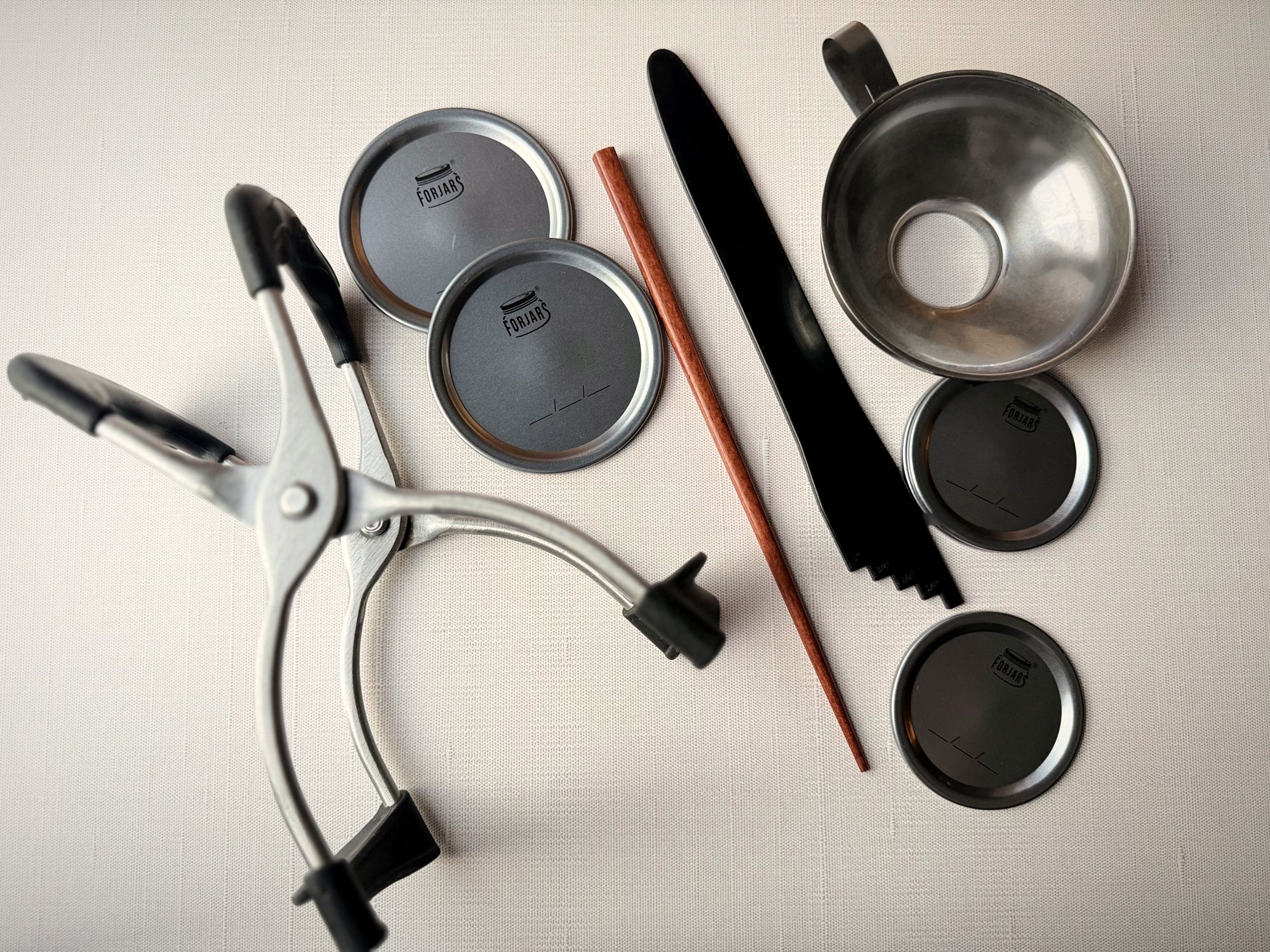 Spring-loaded jar lifter, ForJars canning lids, a wooden bubble stick, a black headspace tool and a stainless steel canning funnel laid out on white linen