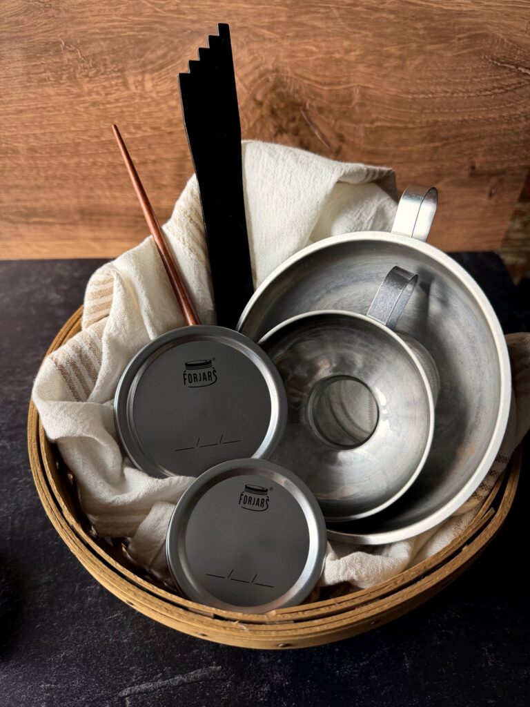Woven basket lined with a cream towel holding two stainless steel canning funnels, ForJars lids, a black headspace tool and a wooden bubble stick