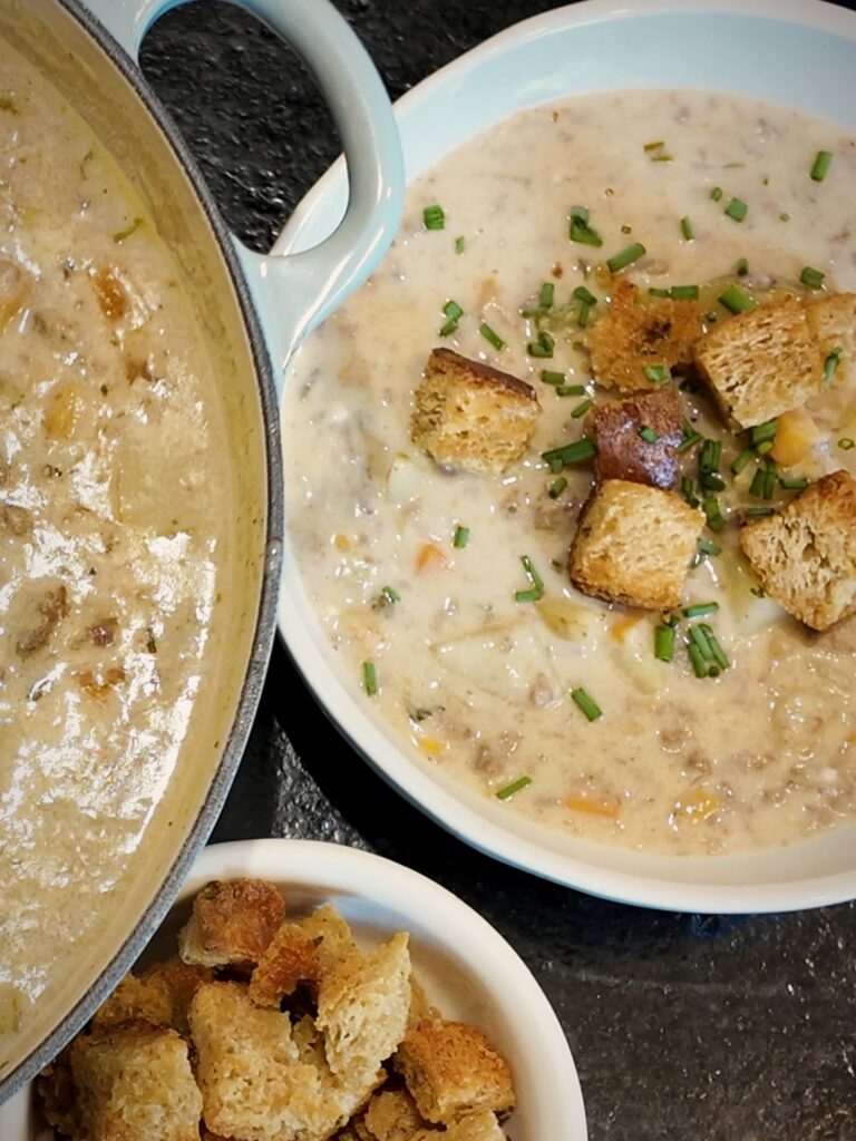 Bowl of cheeseburger soup topped with croutons and chopped chives beside the pot and a small dish of croutons.