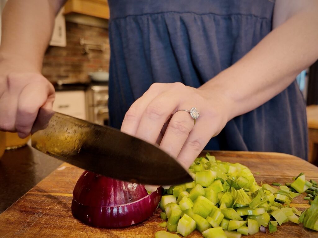 Chef’s knife slicing a red onion on a wooden cutting board next to diced celery.