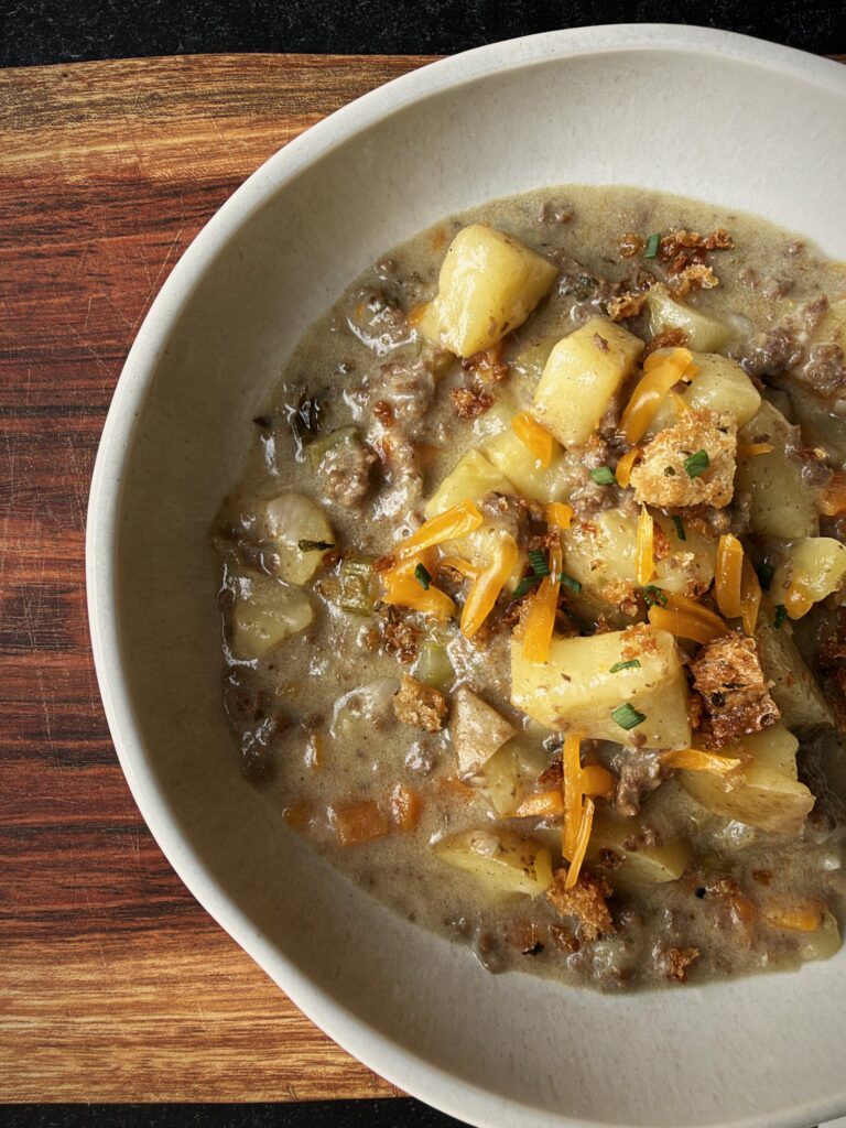 Close-up of creamy cheeseburger soup with potato chunks, ground beef, shredded cheddar, and croutons in a white bowl.