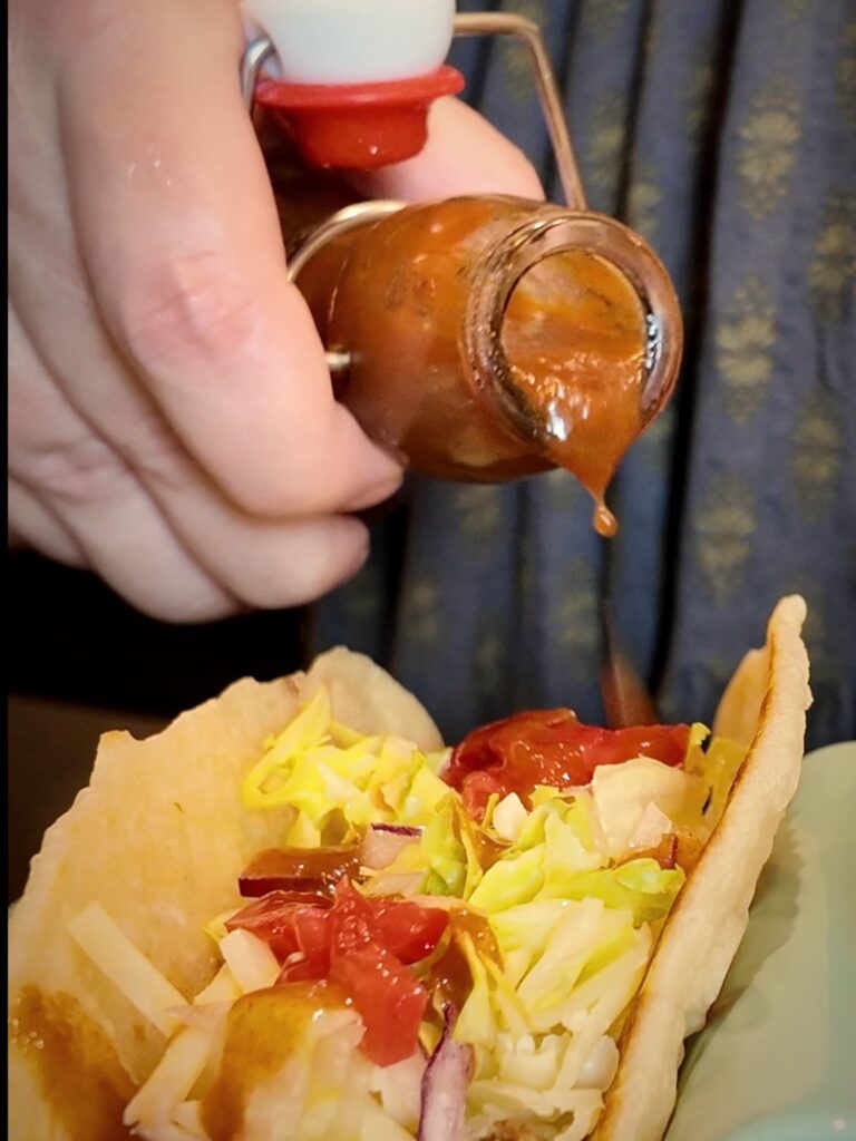 Homemade mild taco sauce pouring from a small glass bottle onto a beef taco with cheese, lettuce and tomatoes