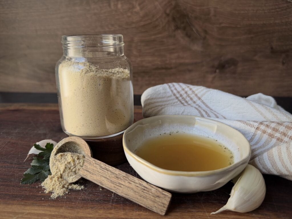 Glass jar of chicken bouillon powder beside a bowl of golden broth, a wooden scoop, garlic and parsley on a wood board