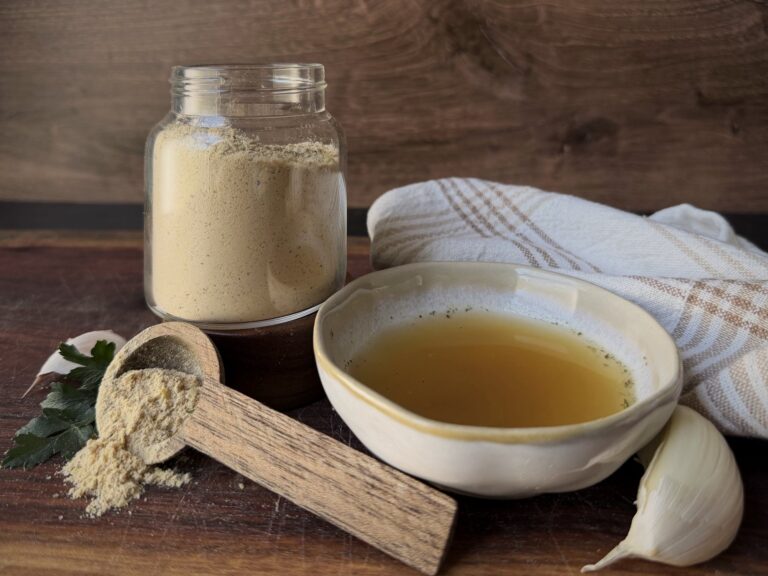 Glass jar of chicken bouillon powder beside a bowl of golden broth, a wooden scoop, garlic and parsley on a wood board
