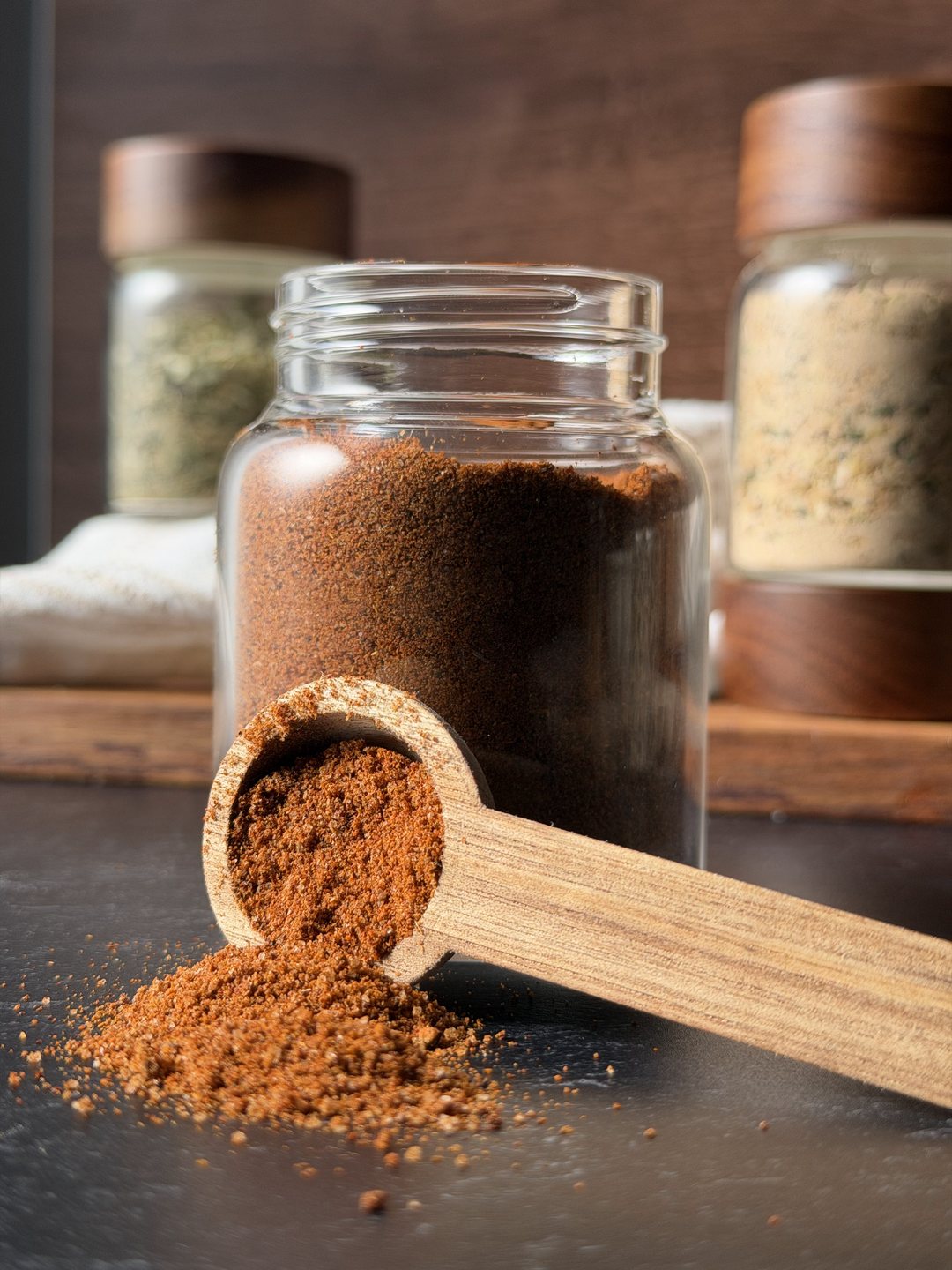 Glass jar of homemade chili seasoning with a wooden scoop spilling seasoning onto a dark counter