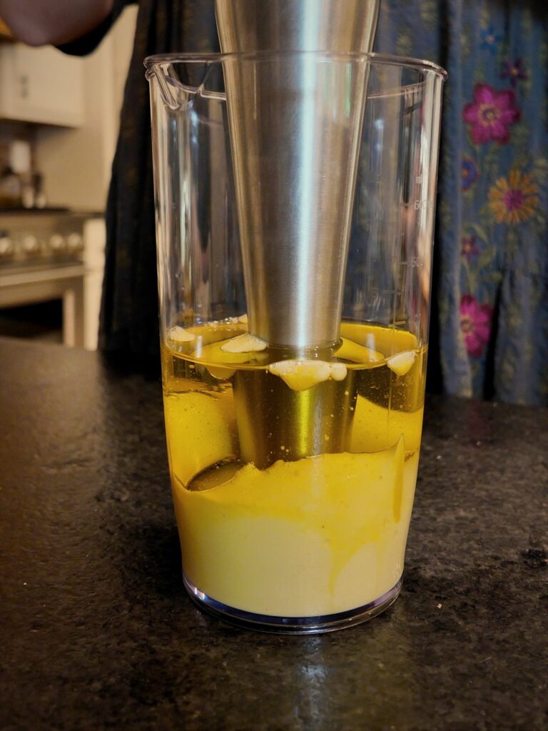 Stainless steel immersion blender in a tall cup, blending oil into homemade mayonnaise.