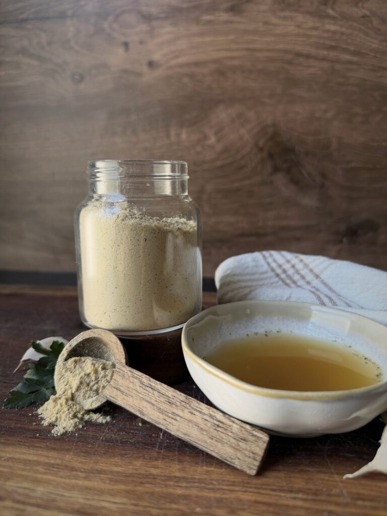 Jar of chicken bouillon powder with a spilled wooden scoop, a bowl of broth and a striped towel against a wood backdrop