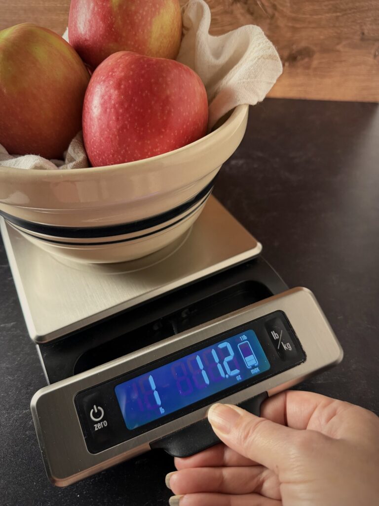 Hand pulling out the display of a stainless digital kitchen scale reading 1 lb 11.2 oz under a stoneware bowl of apples.