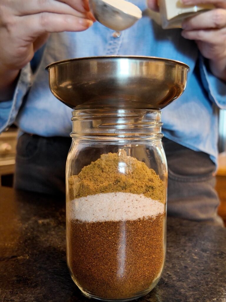 Spices being spooned through a funnel into a mason jar of layered seasoning
