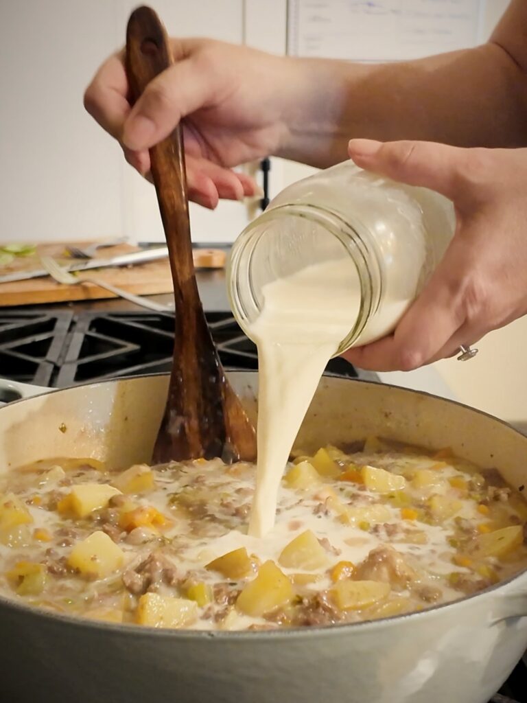 Milk being poured from a glass jar into a pot of cheeseburger soup while a wooden spoon stirs.