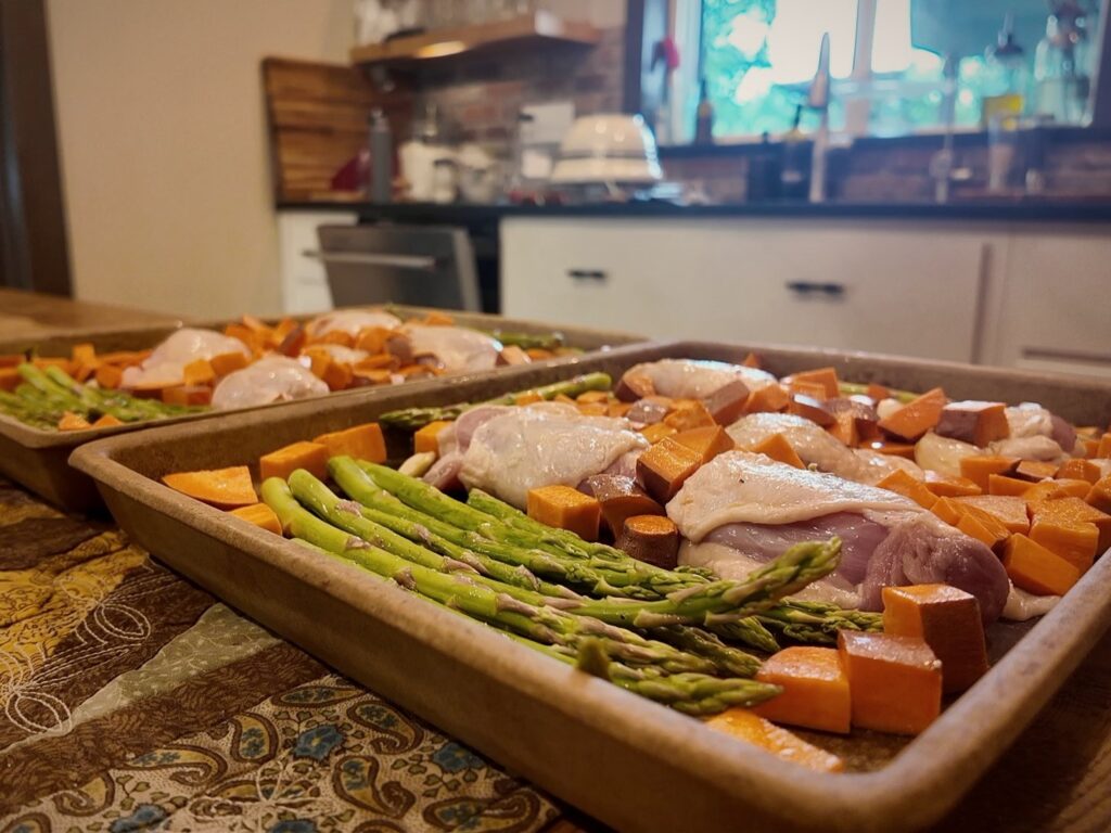 Two rimmed sheet pans of raw chicken thighs, cubed sweet potatoes and asparagus.