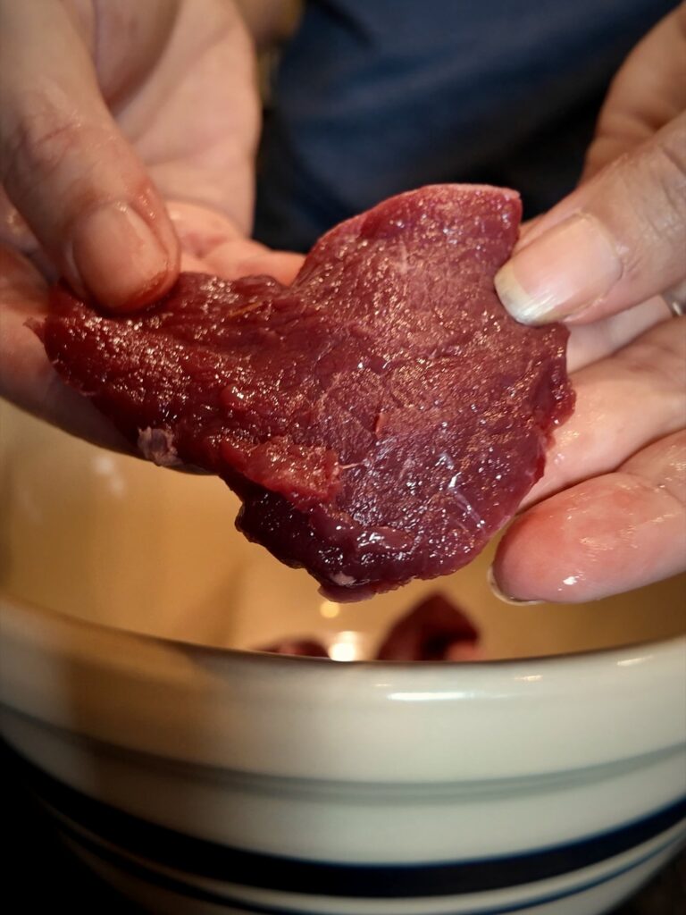 Hands holding a thin slice of raw venison over a blue-striped stoneware bowl