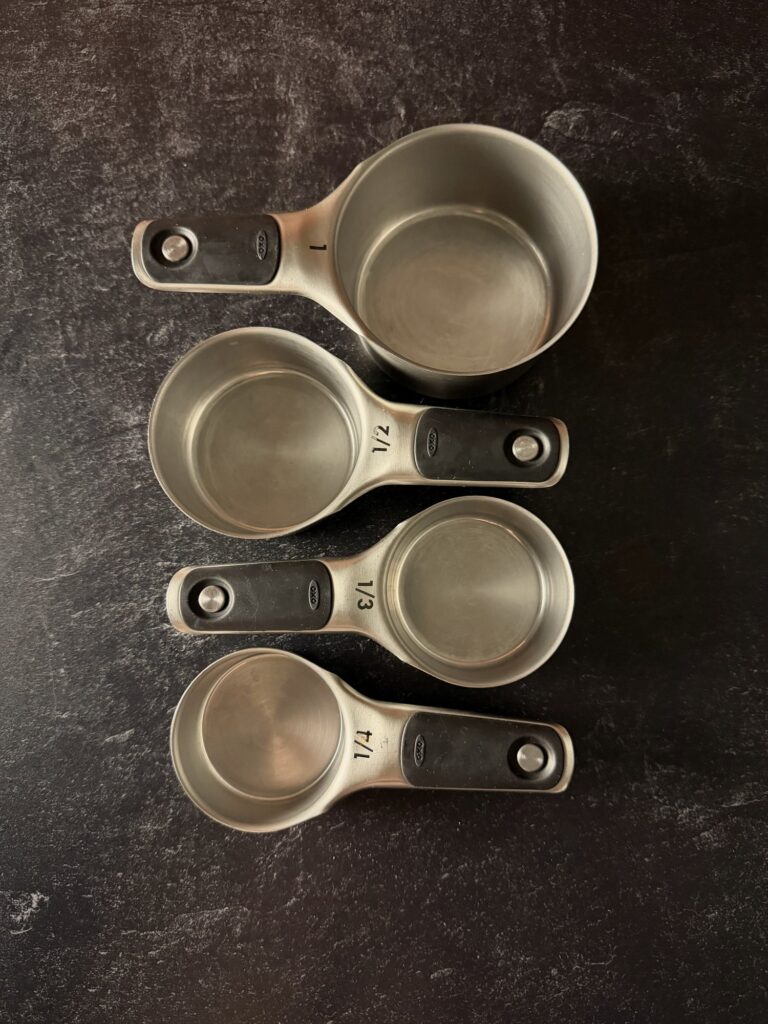 Four stainless steel measuring cups (1, 1/2, 1/3 and 1/4 cup) with black handles on a dark counter.