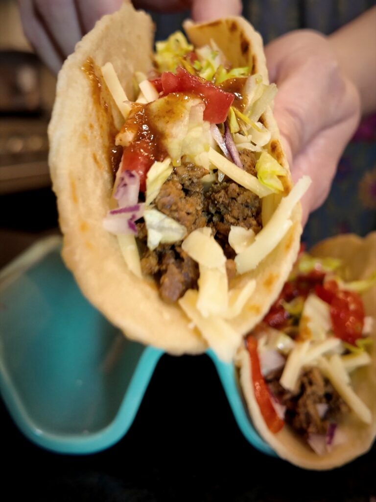 Hand holding a beef taco topped with cheese, cabbage, tomatoes and homemade mild taco sauce