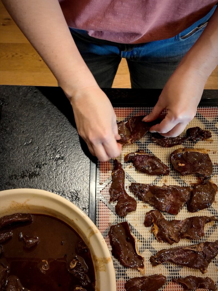Laying marinated venison strips on a dehydrator tray beside a bowl of marinade