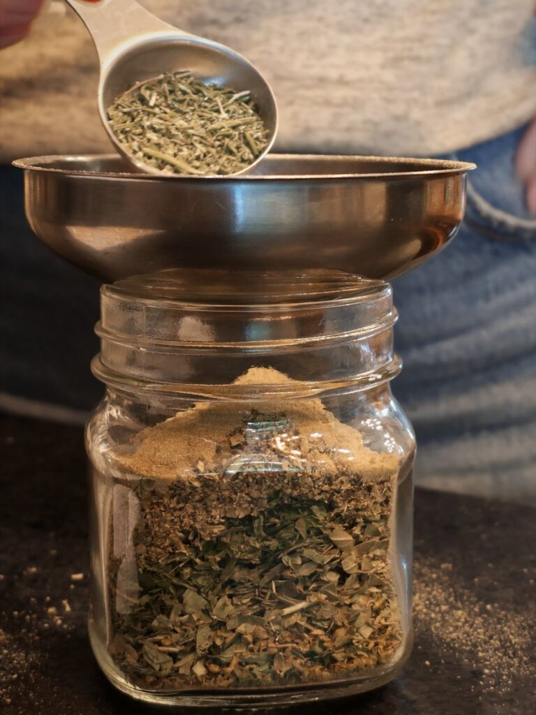 Measuring spoon of dried herbs being added to a glass jar through a stainless steel funnel
