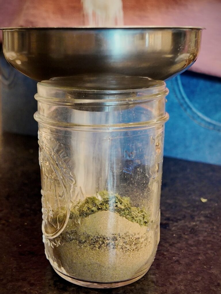 Spooning dried herbs through a stainless steel canning funnel into a glass jar