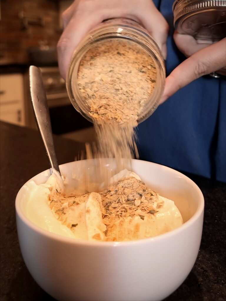 Pouring homemade onion soup mix from a jar onto sour cream in a white bowl