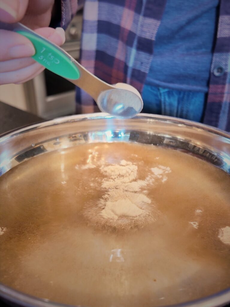 Adding a measuring spoon of white powder to a stainless saucepan of liquid