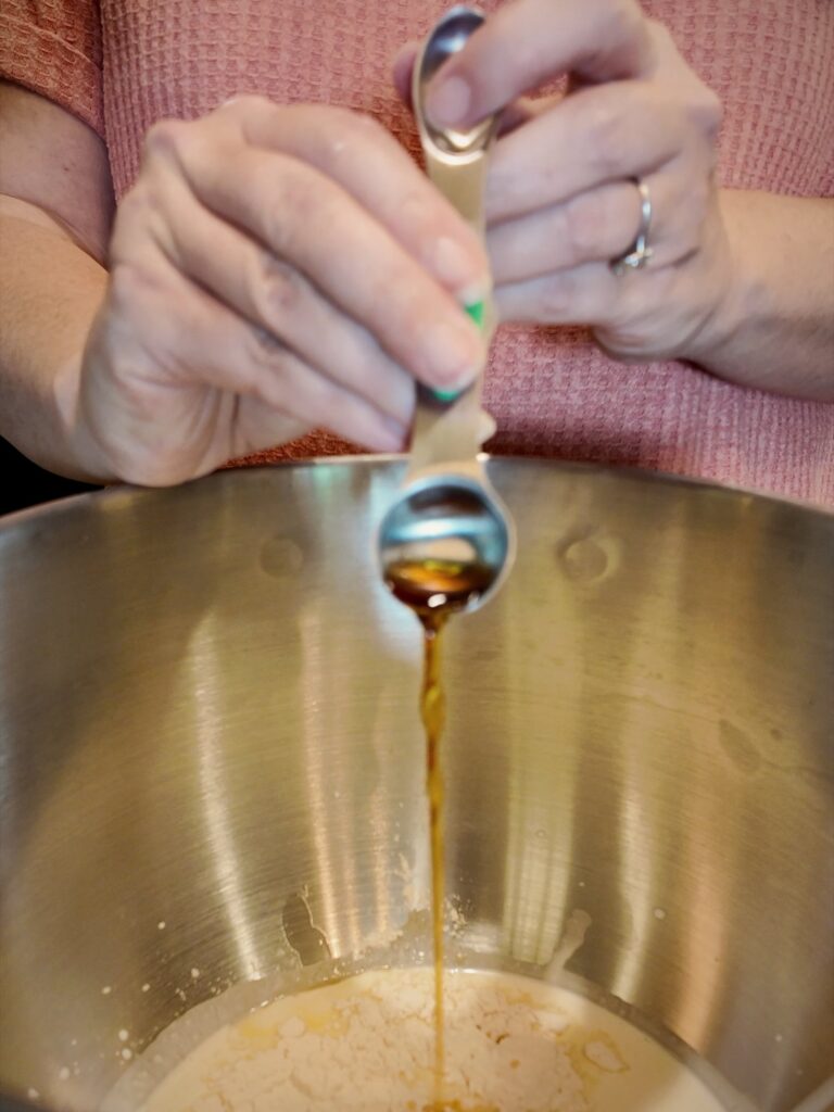 Vanilla extract pouring from a measuring spoon into a mixing bowl of cream