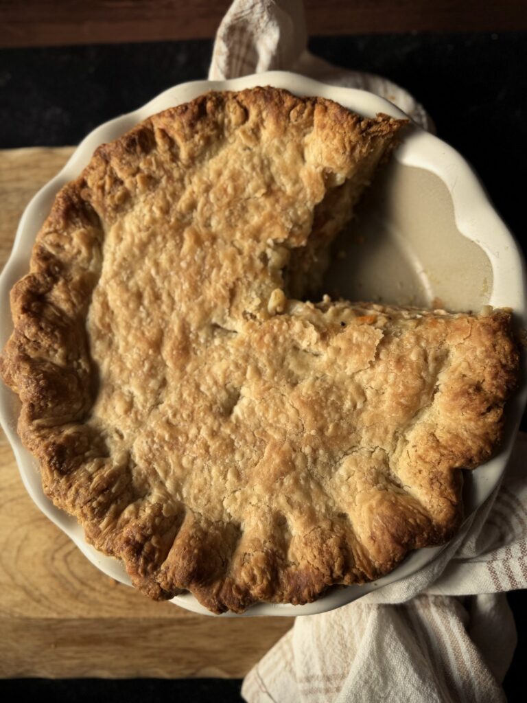 Overhead view of a baked chicken pot pie with one slice removed, in a white fluted pie dish on a wooden board
