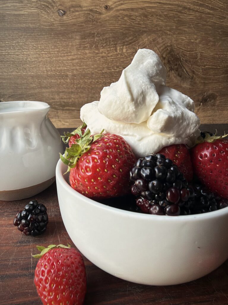 Strawberries and blackberries in a white bowl topped with whipped cream, with loose berries on a wood board