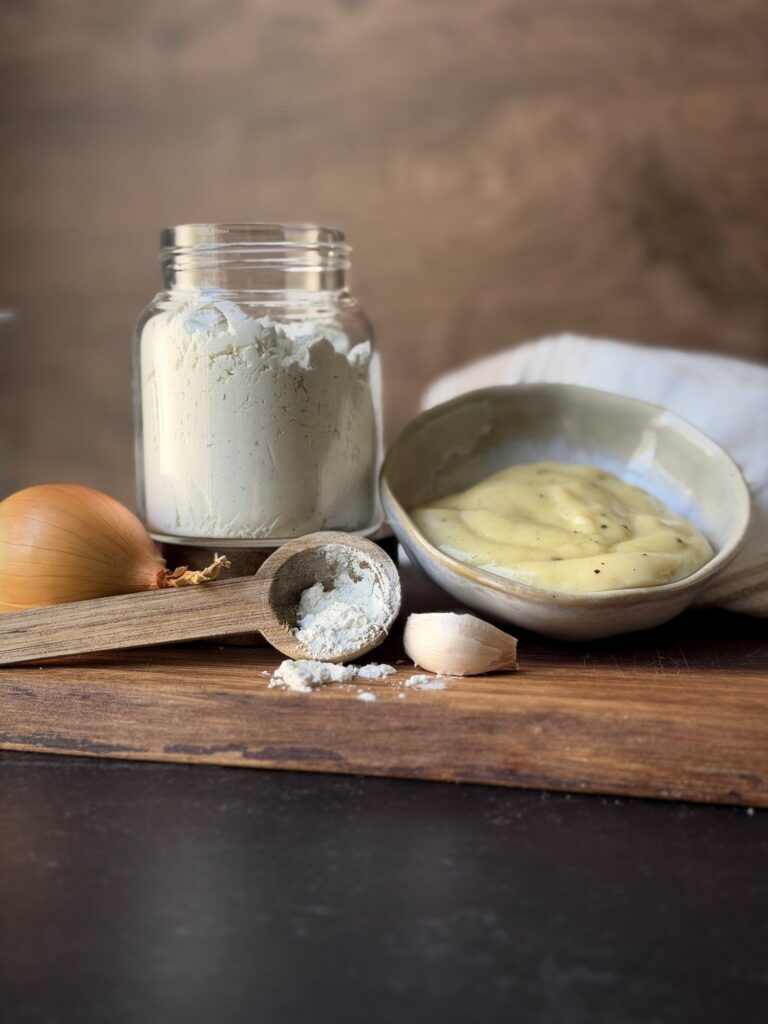 Bowl of thick cream soup beside a jar of dry mix, a wooden scoop, onion and garlic