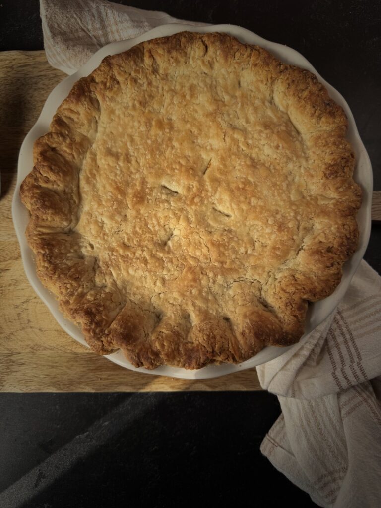 Overhead view of a chicken pot pie with a golden all-butter top crust and crimped edges in a white fluted pie dish on a wooden board