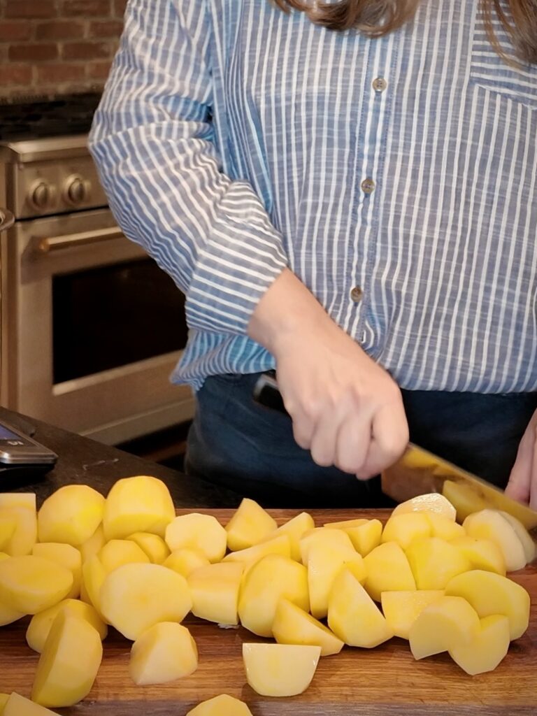 Woman in a striped shirt chopping peeled Yukon Gold potatoes into chunks on a wooden cutting board.