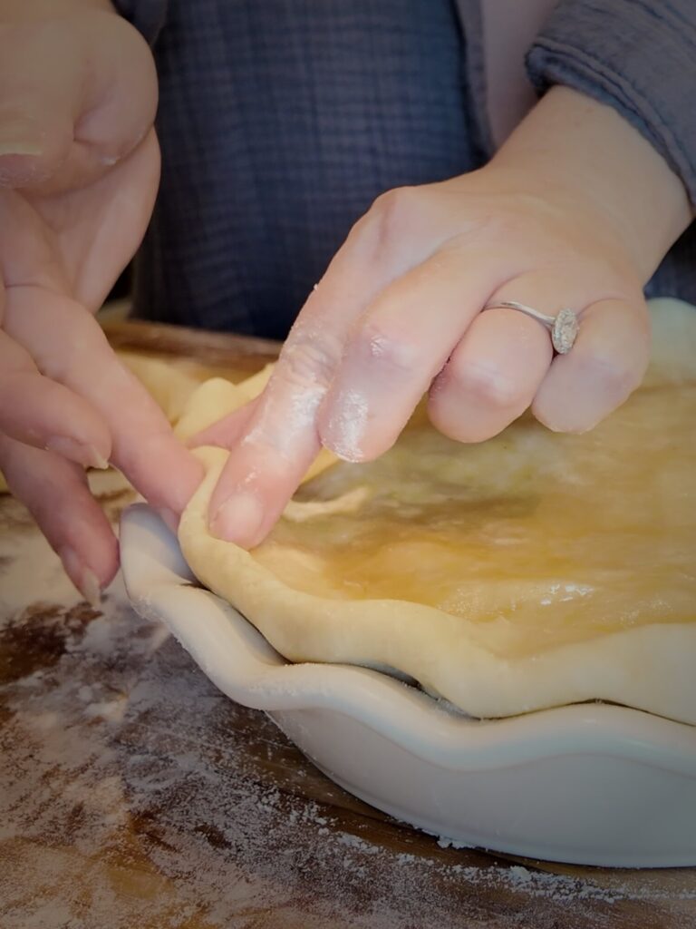Fingers pinching the edge of an unbaked top crust on a pie in a white fluted pie dish