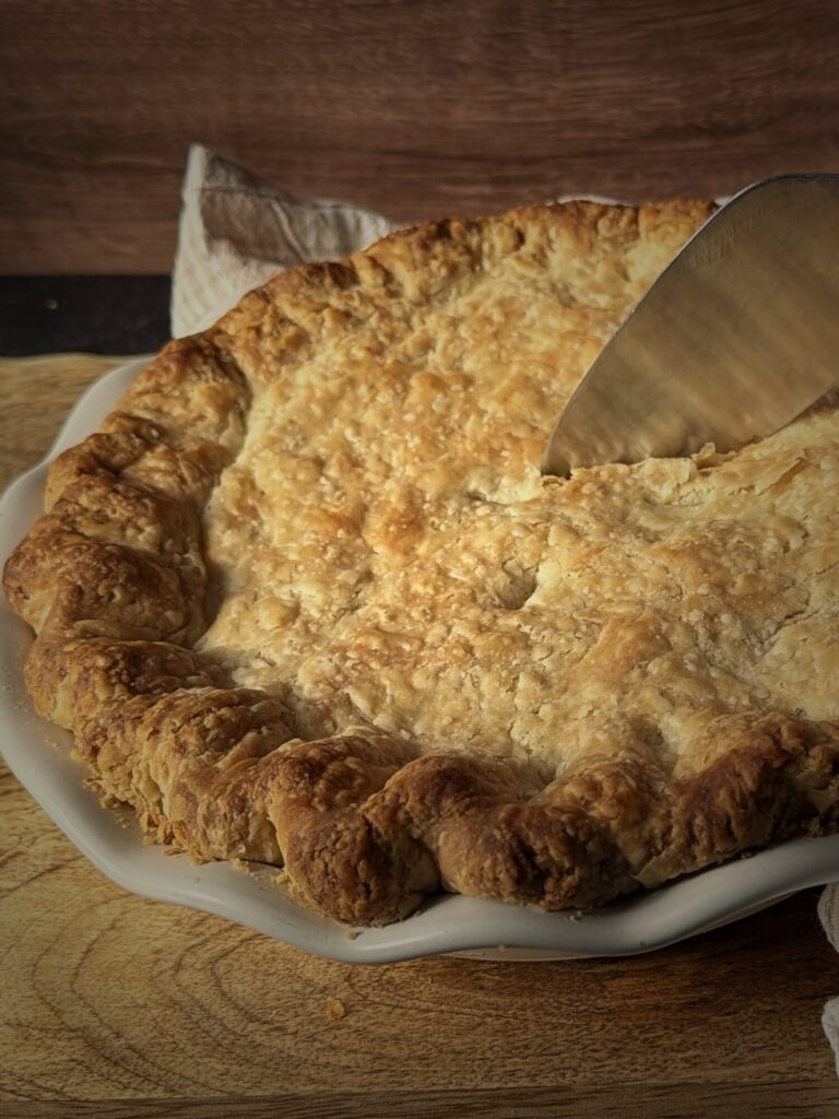 Pie server cutting into the flaky top crust of a chicken pot pie in a white fluted pie dish