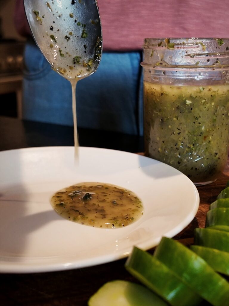 Homemade Italian dressing dripping from a spoon onto a white plate, with a jar of dressing and sliced cucumbers