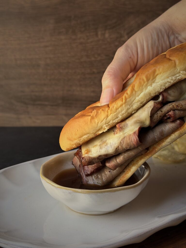 Hand dunking a roast beef and provolone French dip sandwich into a bowl of au jus